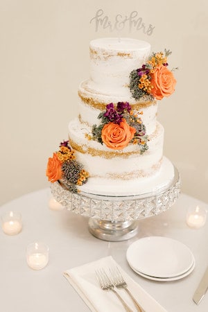 three tiered naked wedding cake with purple and orange flowers sitting on a crystal cake stand