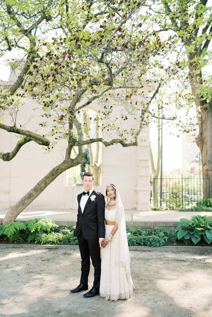 Bride and groom stand under large trees at the Rodin Museum wedding venue